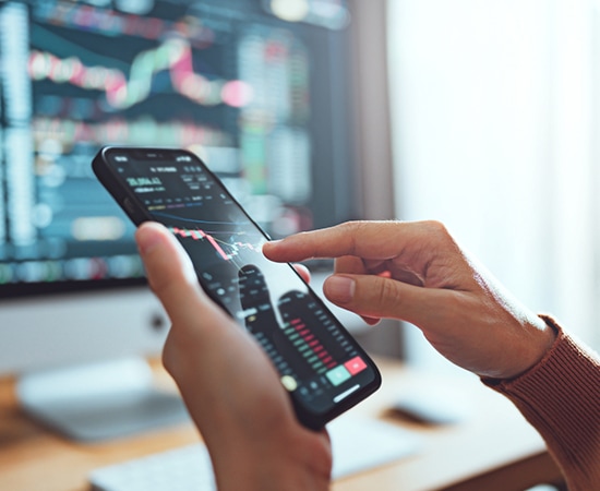 A person using a smartphone to analyze stock market data is shown. The phone screen displays a graph with red and green candlesticks. In the background, an Acadian computer setup showcases various financial charts and information.