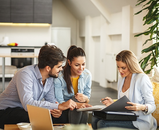 Three people are sitting around a table in a modern living room. A woman on the right is holding a folder and a pen, explaining real estate documents to a man and another woman on the left. There is a laptop on the table, and the atmosphere is collaborative.