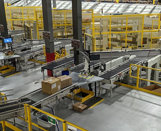 A warehouse with conveyor belts transporting packages. The area is equipped with various machinery and material handling equipment, including Dematic robotic arms for handling items. Yellow railings and safety barriers are present, creating an organized and automated space.