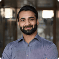 A man with a beard and dark hair is smiling and facing the camera. He is wearing a light purple button-up shirt, standing in front of a blurred background featuring a wooden frame. The well-lit, professional atmosphere makes his employee bio picture stand out beautifully.