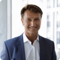 A smiling man with short, light brown hair wearing a light blue suit jacket and white dress shirt stands indoors in front of a window, where the blurred cityscape resembles colorful blocks in the background.
