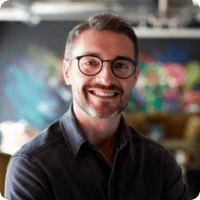 A smiling man with short hair, glasses, and a beard is wearing a dark shirt. He is standing indoors against a colorful, blurred background often seen in employee grid bios.