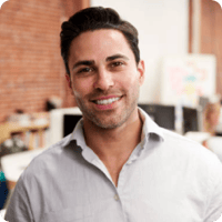 A man with dark hair and a neatly trimmed beard is standing in an office, smiling at the camera. He is wearing a light-colored button-up shirt. The background features a brick wall, a desk, and some office equipment. This photo is part of our Employee Bios that showcase our dedicated team members.