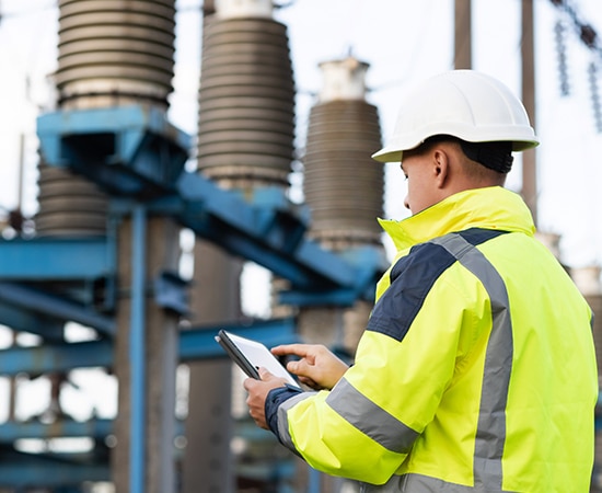 A worker in a bright yellow safety jacket and white hard hat is standing outdoors at an industrial site, holding and using a tablet. Large cylindrical equipment and metal structures, part of the ABB Power Systems division, are visible in the background.