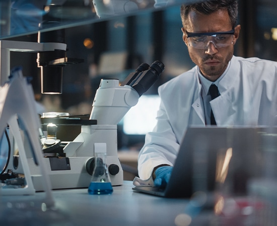 A scientist in a white lab coat and protective eyewear works in a Rockstep Solutions-equipped laboratory. He is using a laptop and is seated beside a microscope and various scientific equipment. The laboratory is well-lit with a modern design.