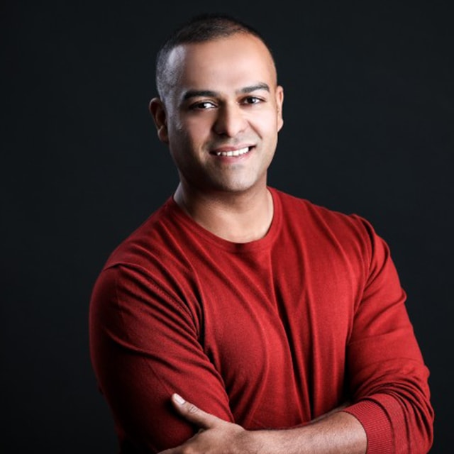 A person with a close-cropped haircut smiles at the camera against a dark background. They are wearing a long-sleeved red shirt and have their arms folded across their chest, exuding confidence and a strong sense of culture.