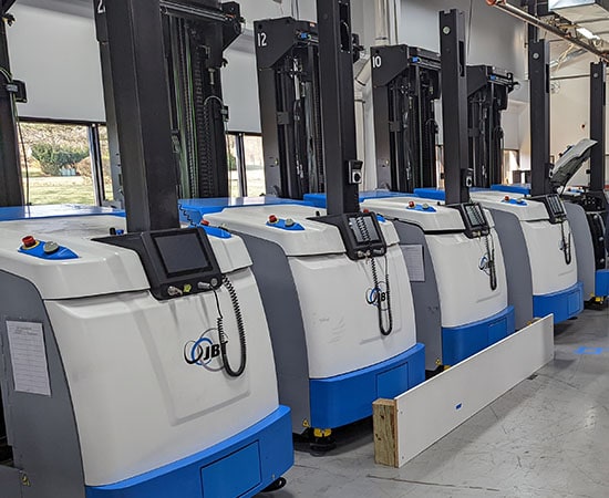A row of white and blue automated guided vehicles (AGVs) with control panels and screens are lined up in an industrial warehouse setting. The AGVs are marked with numbers, positioned under tall, black lifting mechanisms, and seamlessly integrated with cloud migration systems. The floor is clean and well-kept.