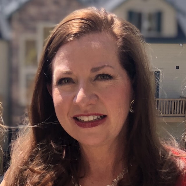 A woman with long brown hair and a warm smile is pictured outdoors. She is wearing earrings and a necklace that reflect the richness of culture, and the background features a blurred view of a building.