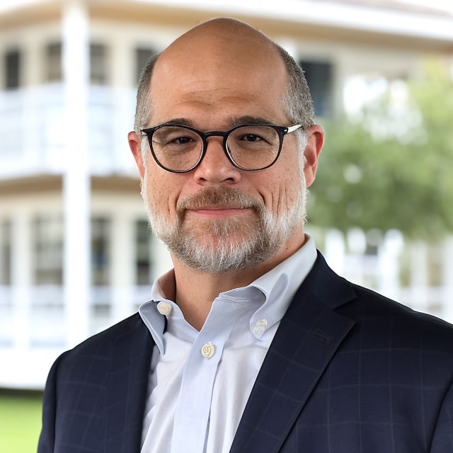A man with short hair, glasses, and a beard is wearing a dark suit jacket over a light blue dress shirt. He is standing outdoors in front of a blurred background that captures the essence of urban culture, featuring a white building with multiple floors and windows.
