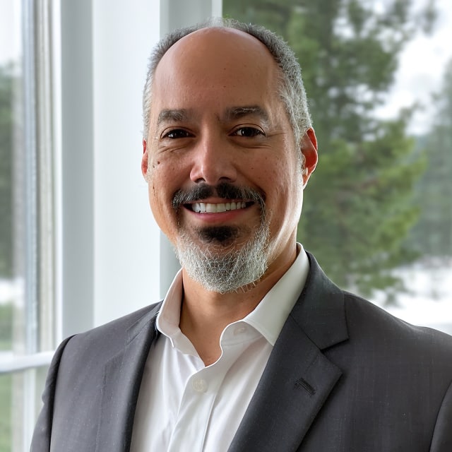 A man with a salt-and-pepper beard and mustache, wearing a suit and white dress shirt, is smiling at the camera. He is standing indoors with large windows behind him, showcasing a view of greenery outside. His warm smile reflects the rich culture he proudly embodies.