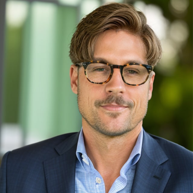 A man with light brown hair, wearing glasses and a navy blue suit over a light blue shirt, stands in front of a blurred outdoor background featuring greenery. He is smiling slightly and looks composed, exuding an air of cultured sophistication.
