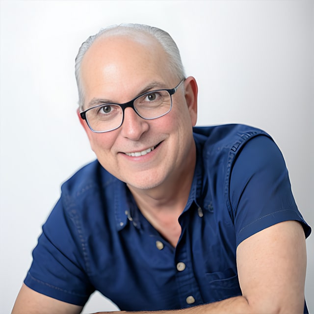 A smiling older man with glasses, short white hair, and a blue button-up shirt poses against a plain white background. Leaning slightly forward, he exudes a friendly demeanor that radiates warmth and culture.