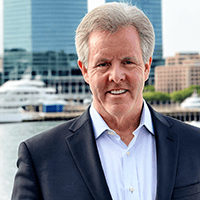A man with gray hair, wearing a dark suit and white shirt, smiles in front of a waterfront backdrop, featuring large boats and modern buildings that reflect the rich culture of the city.