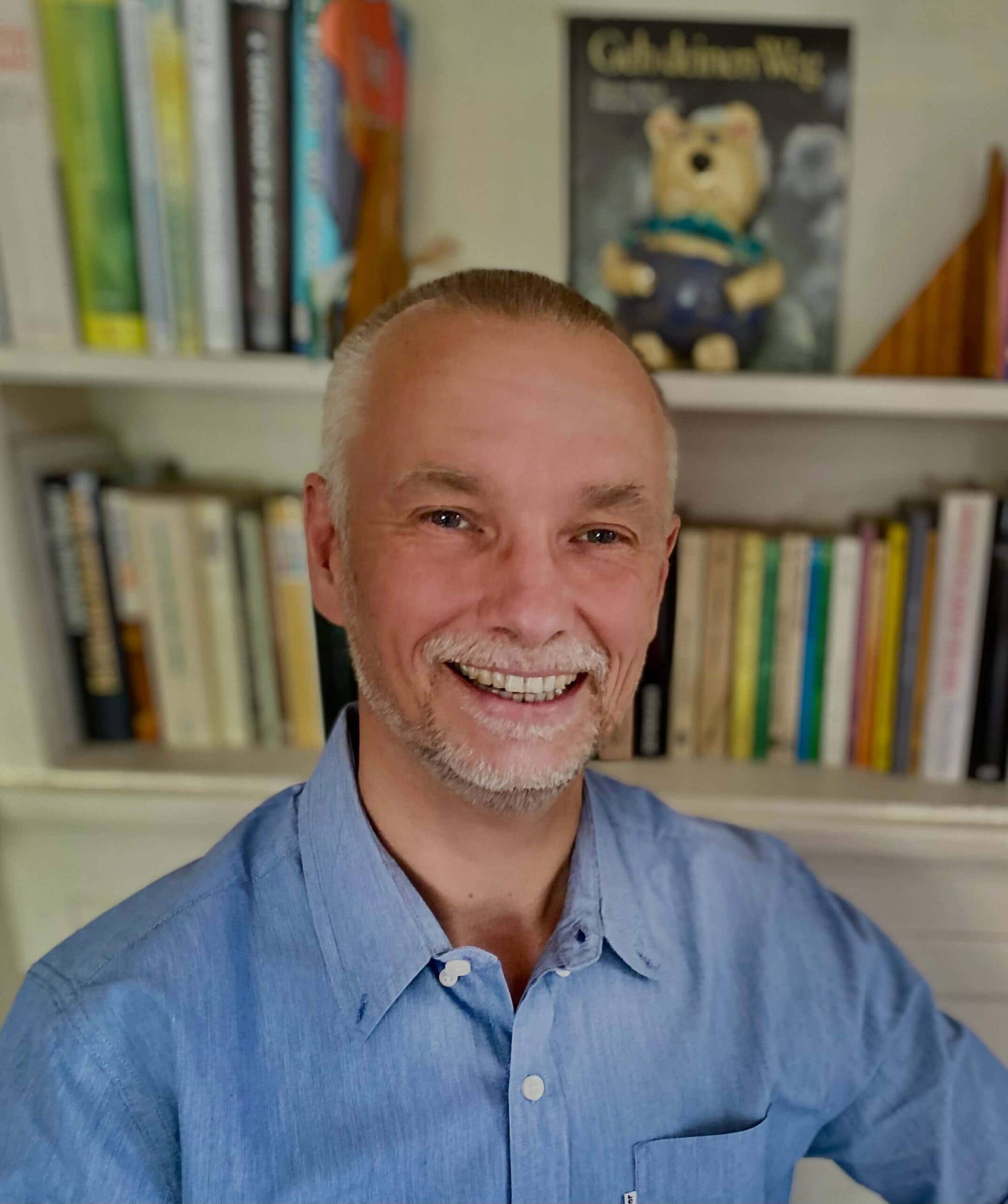 A smiling man with a closely trimmed beard and short, light hair is wearing a blue shirt. He is standing in front of a bookshelf filled with various books that reflect diverse cultures and a small plush bear.