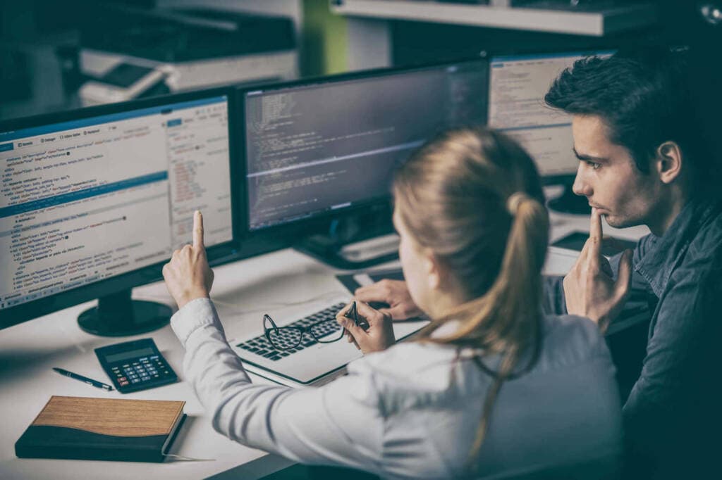 Two people working at a desk with three computer monitors displaying code. The person on the left is pointing at one of the screens related to DevOps, while the person on the right has a thoughtful expression, touching their face. A calculator and a notepad are on the desk for monitoring tasks.