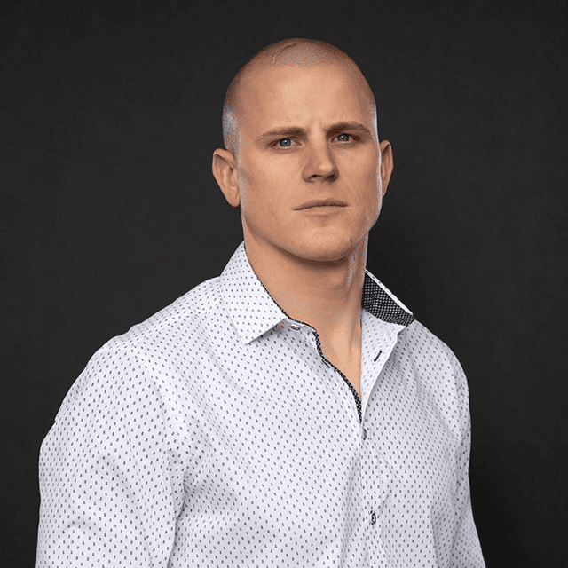 A person wearing a white patterned shirt is looking directly at the camera against a dark background.