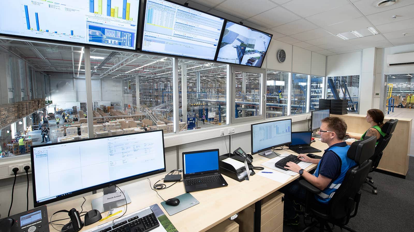 Two people monitor computer screens in a Dematic Control Tower, overseeing a busy warehouse floor filled with shelves and workers.