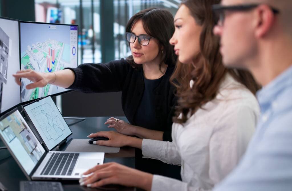 Three cloud engineers collaborate at computers, reviewing geospatial architectural designs and building plans on screens and laptops.